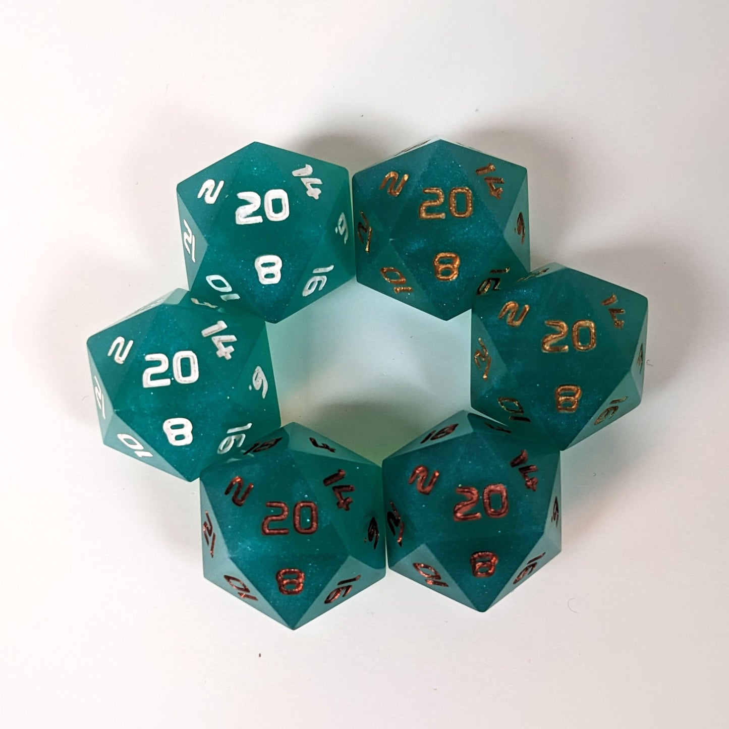 Three pairs of standard d20s are arranged in a hexagon on a white background. All of the handmade dice are teal with a pearlescent shine. The two d20s on the top right have gold numbers, the two d20s on the top left have white numbers, and the bottom two have copper numbers, showing how each number color affects the appearance of the hue of the pearlescent teal dice.