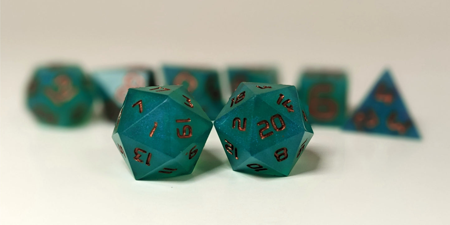 A straight on photo of a standard set of DnD dice that are teal with a pearlescent shine with copper numbers focusing on 2 d20s in the center. The d20 on the left has the 1 face centered and the d20 on the right has the 20 face centered. The other 6 dice are lined up in the background but are out of focus, while the d20s are in sharp focus.