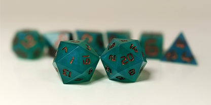 A straight on photo of a standard set of DnD dice that are teal with a pearlescent shine with copper numbers focusing on 2 d20s in the center. The d20 on the left has the 1 face centered and the d20 on the right has the 20 face centered. The other 6 dice are lined up in the background but are out of focus, while the d20s are in sharp focus.