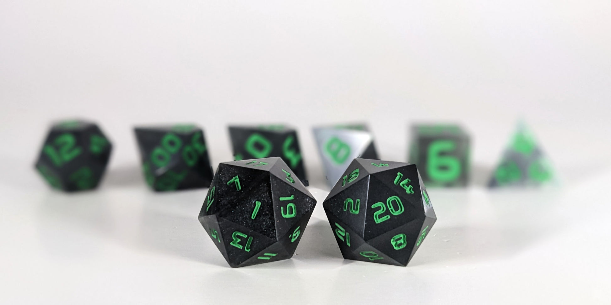 A straight on photo of a standard set of dice that are black with some semitranslucent swirls and bright green numbers focusing on 2 d20s in the center. The d20 on the left has the 1 face centered and the d20 on the right has the 20 face centered. The other 6 dice are lined up in the background but are out of focus, while the d20s are in sharp focus.