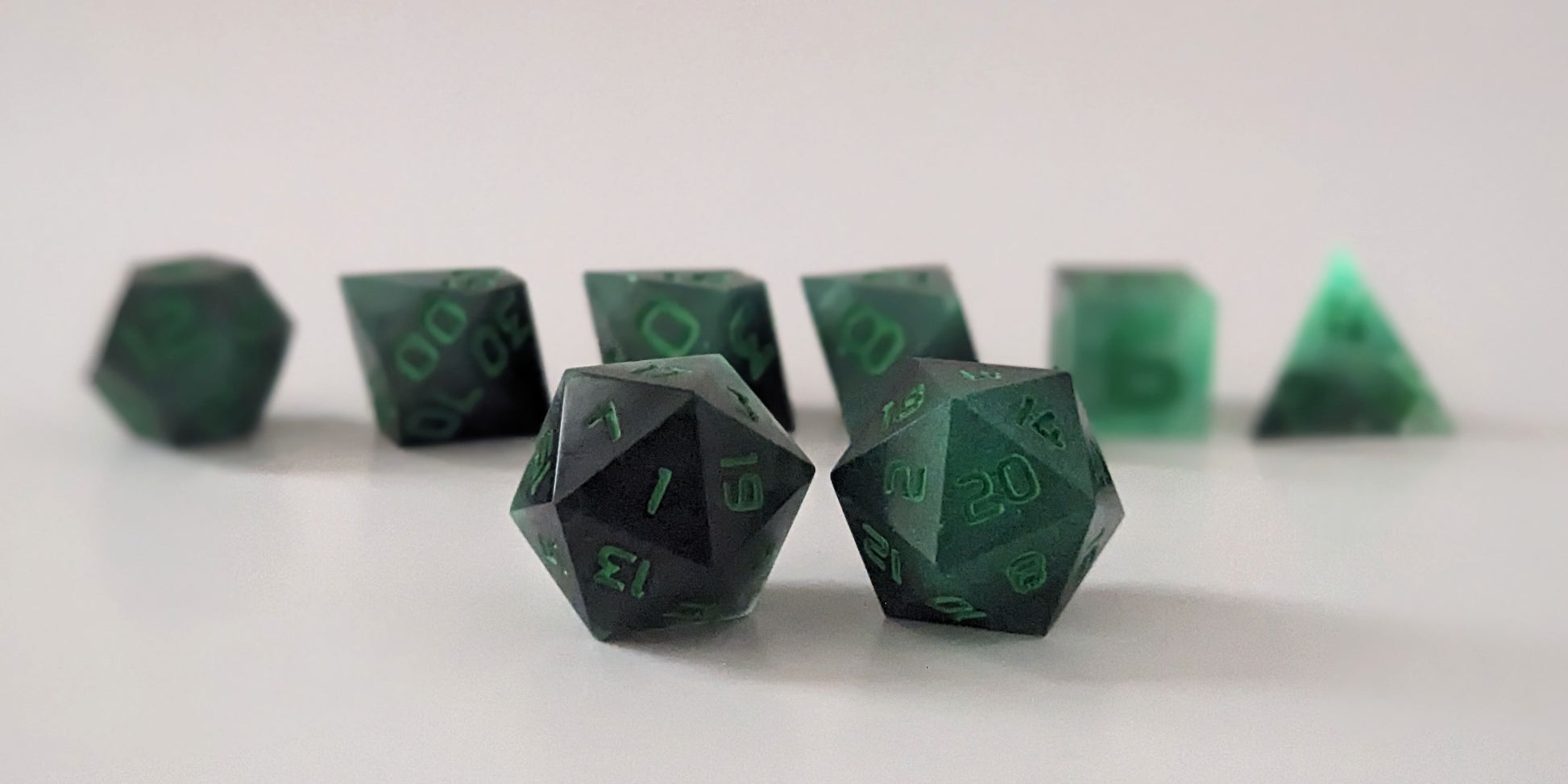 A straight on photo of a standard set of dice that are glowing green in the dark with the black still visible and green numbers focusing on 2 d20s in the center. The d20 on the left has the 1 face centered and the d20 on the right has the 20 face centered. The other 6 dice are lined up in the background but are out of focus, while the d20s are in sharp focus.