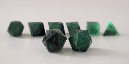 A straight on photo of a standard set of dice that are glowing green in the dark with the black still visible and green numbers focusing on 2 d20s in the center. The d20 on the left has the 1 face centered and the d20 on the right has the 20 face centered. The other 6 dice are lined up in the background but are out of focus, while the d20s are in sharp focus.