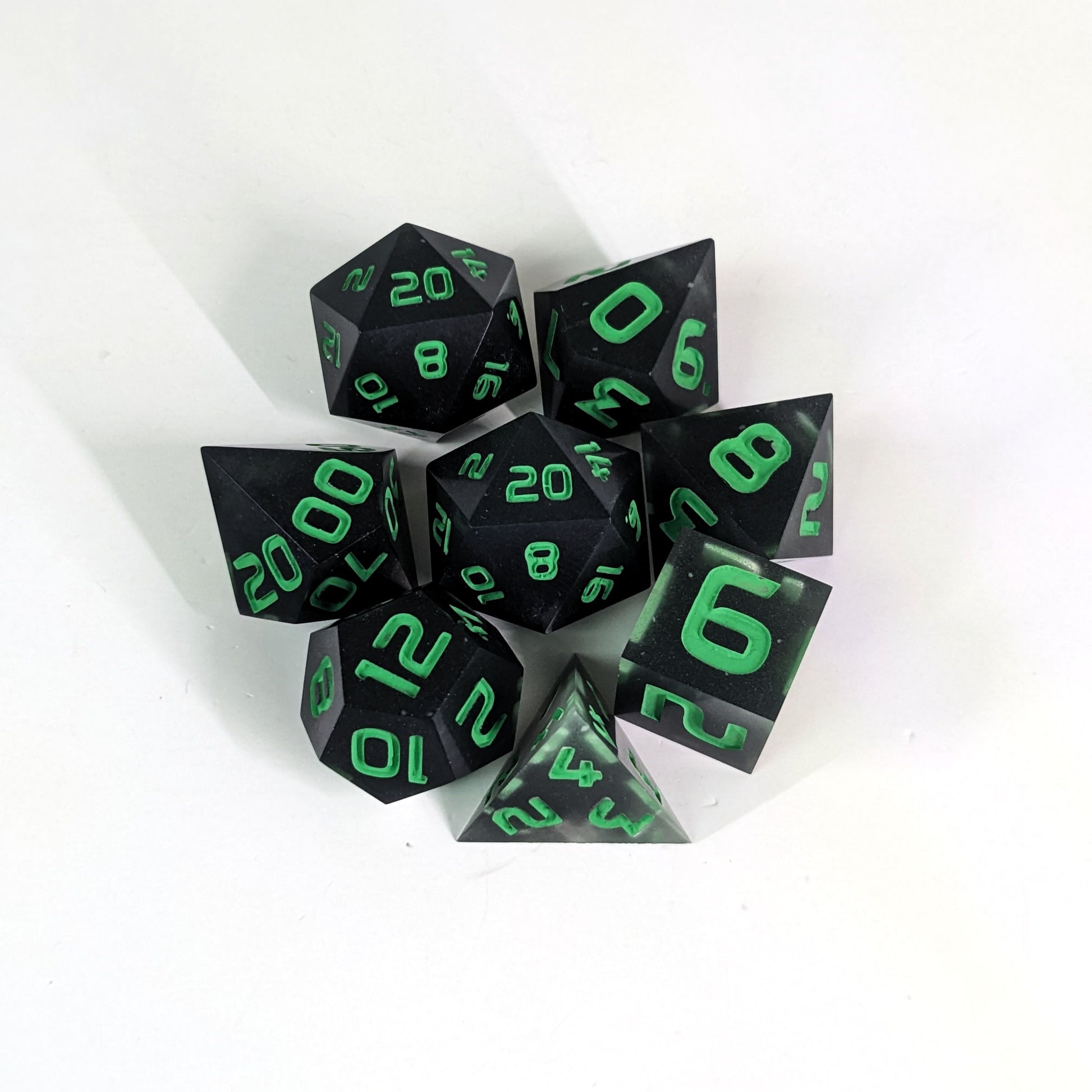 A complete set of 8 handmade artisan dice that are black with some semitranslucent swirls and bright green numbers. The DnD dice set is arranged in a circle and features 2 D20s, one of which is in the center surrounded by the other 7 dice.