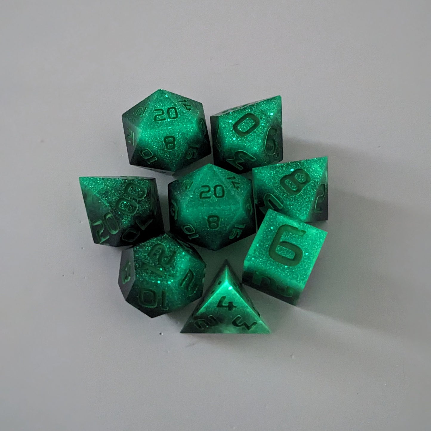 A complete set of 8 handmade artisan dice that are glowing green in the dark with the black still visible and green numbers. The DnD dice set is arranged in a circle and features 2 D20s, one of which is in the center surrounded by the other 7 dice.