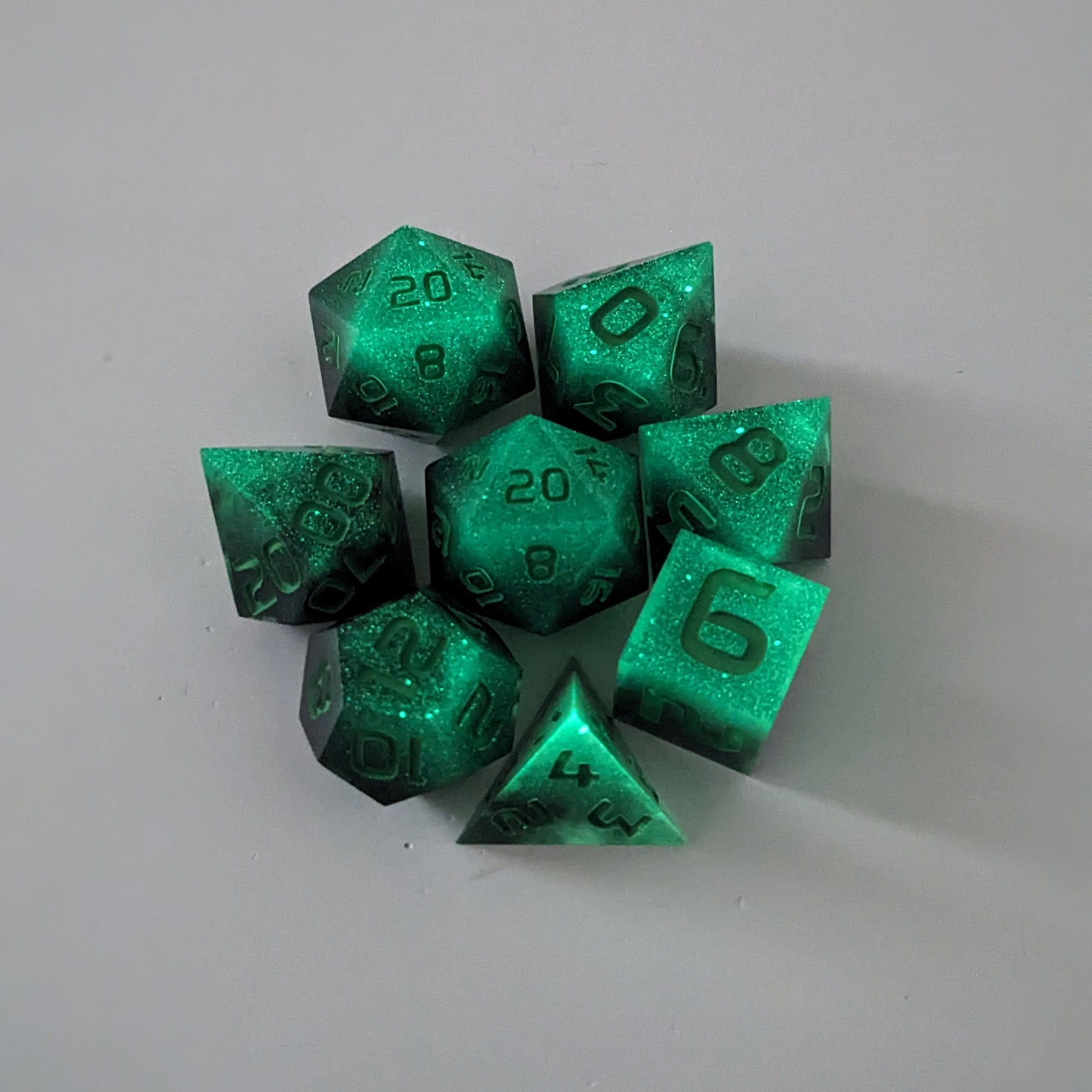 A complete set of 8 handmade artisan dice that are glowing green in the dark with the black still visible and green numbers. The DnD dice set is arranged in a circle and features 2 D20s, one of which is in the center surrounded by the other 7 dice.