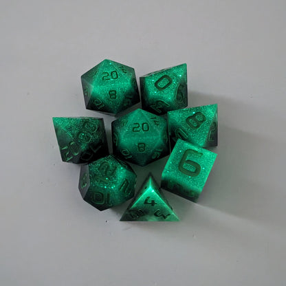 A complete set of 8 handmade artisan dice that are glowing green in the dark with the black still visible and green numbers. The DnD dice set is arranged in a circle and features 2 D20s, one of which is in the center surrounded by the other 7 dice.