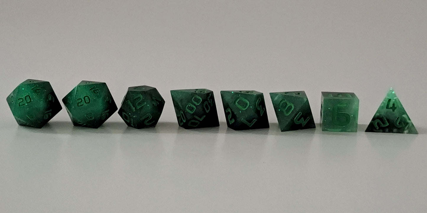 A straight on photo of a complete set of polyhedral dice that are glowing green in the dark with the black still visible and green numbers arranged in a straight line. They are lined up with the 2 d20s on the left going down to the d4 on the right side. The highest value number of each die is facing forward.