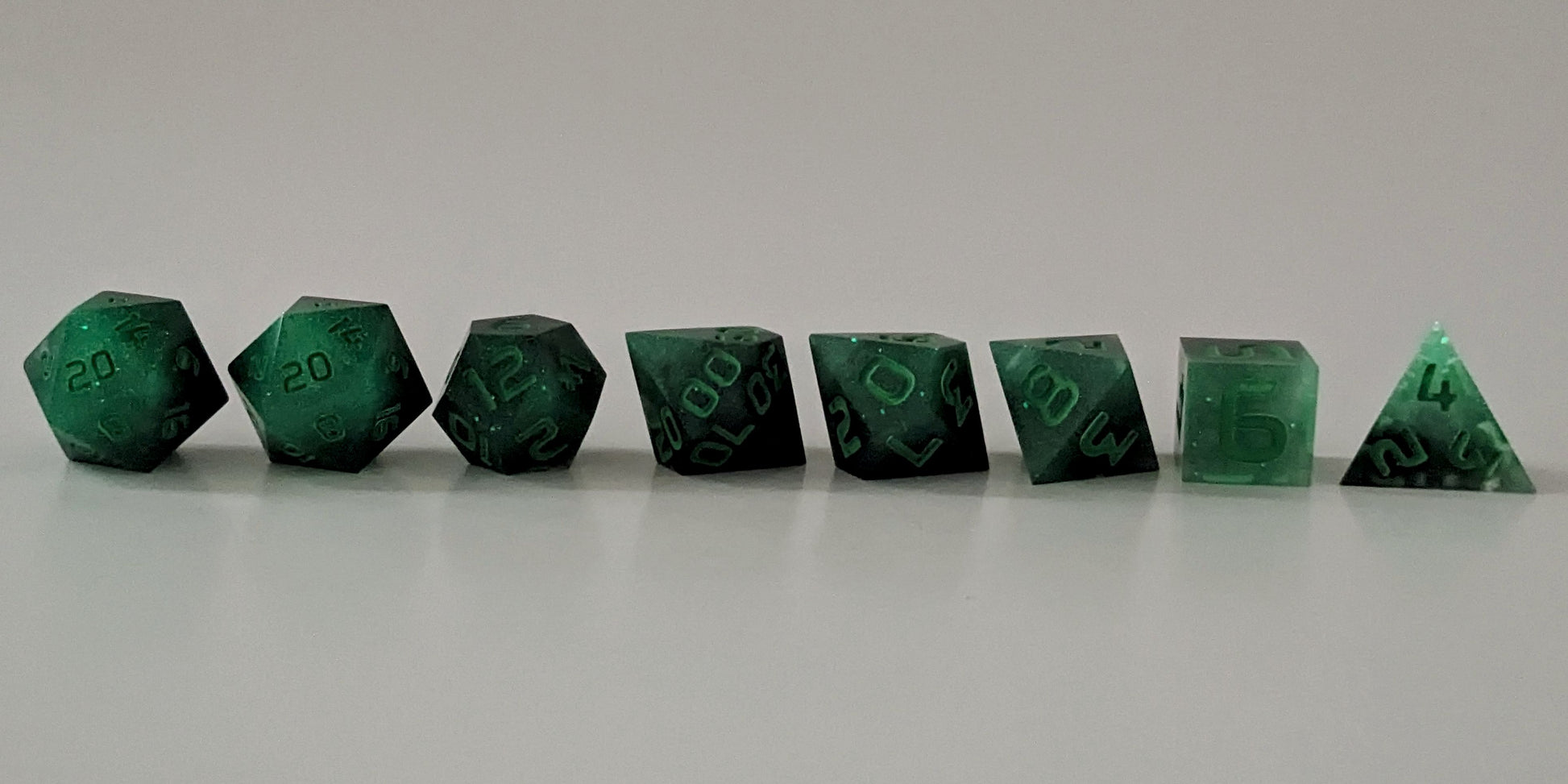 A straight on photo of a complete set of polyhedral dice that are glowing green in the dark with the black still visible and green numbers arranged in a straight line. They are lined up with the 2 d20s on the left going down to the d4 on the right side. The highest value number of each die is facing forward.