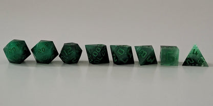 A straight on photo of a complete set of polyhedral dice that are glowing green in the dark with the black still visible and green numbers arranged in a straight line. They are lined up with the 2 d20s on the left going down to the d4 on the right side. The highest value number of each die is facing forward.