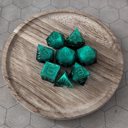 A complete set of 8 handmade artisan dice that are glowing green in the dark with the black still visible and green numbers. The DnD dice set is on a wooded circle and the dice are arranged in a circle and features 2 D20s, one of which is in the center surrounded by the other 7 dice.