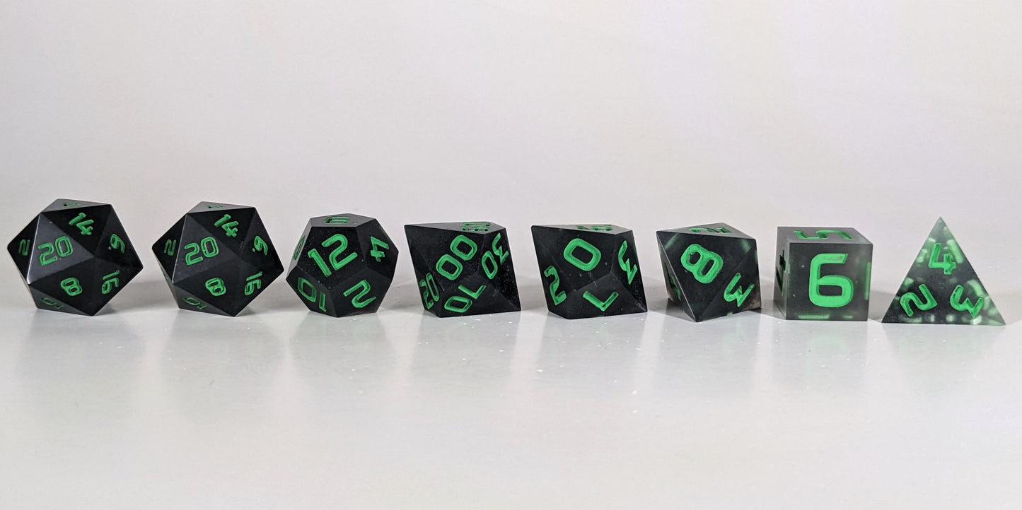 A straight on photo of a complete set of polyhedral dice that are black with some semitranslucent swirls and bright green numbers arranged in a straight line. They are lined up with the 2 d20s on the left going down to the d4 on the right side. The highest value number of each die is facing forward.