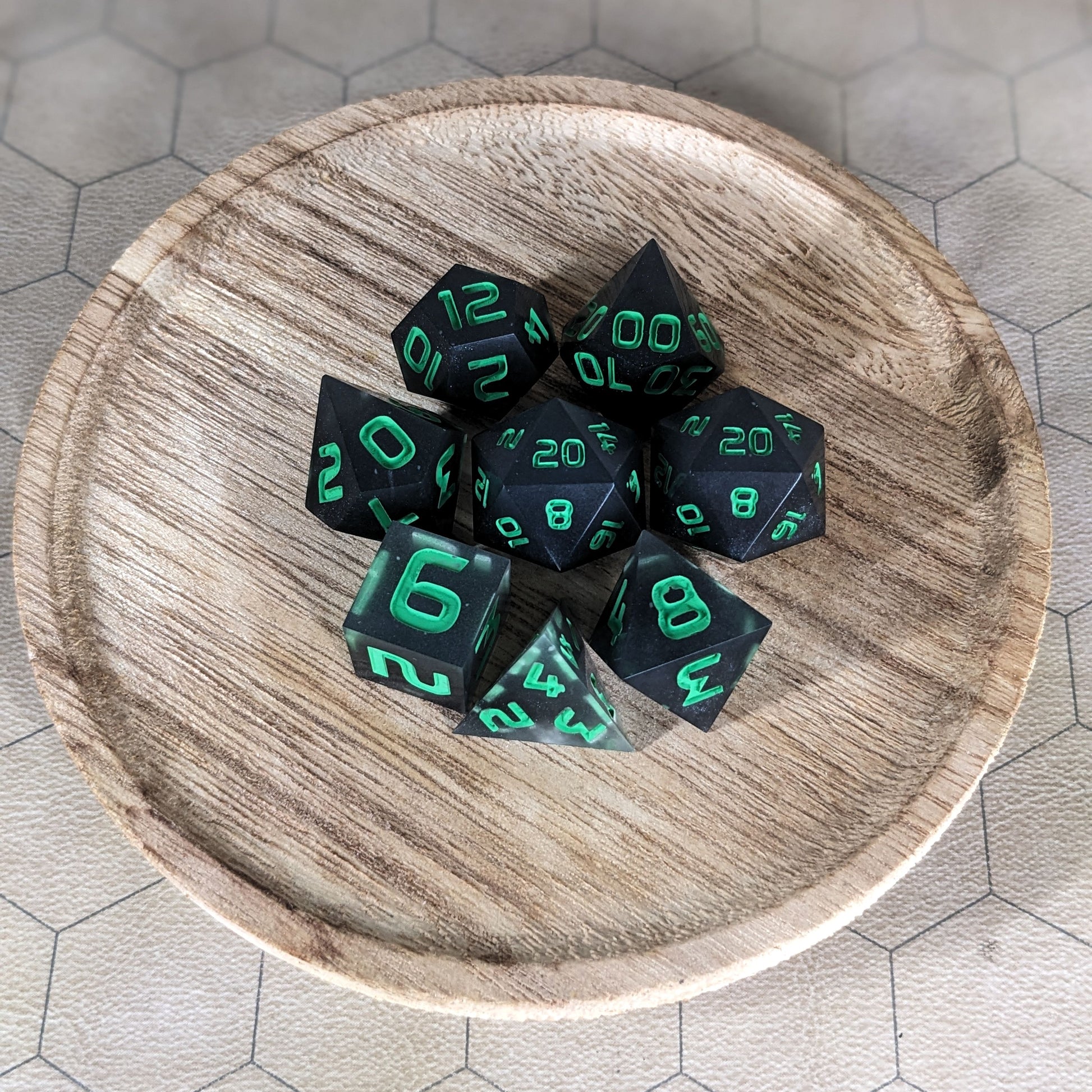 A complete set of 8 handmade artisan dice that are black with some semitranslucent swirls and bright green numbers. The DnD dice set is on a wooded circle and the dice are arranged in a circle and features 2 D20s, one of which is in the center surrounded by the other 7 dice.