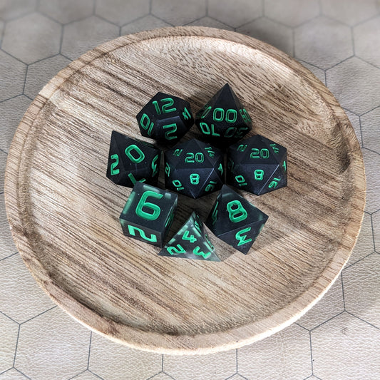 A complete set of 8 handmade artisan dice that are black with some semitranslucent swirls and bright green numbers. The DnD dice set is on a wooded circle and the dice are arranged in a circle and features 2 D20s, one of which is in the center surrounded by the other 7 dice.
