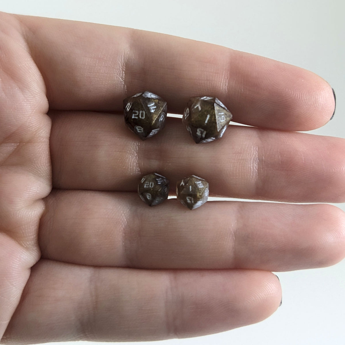 Two pairs of handmade d20 earrings with titanium hardware that are brown and yellow tiger's eye swirls with white numbers held between the fingers of a flat hand. The larger pair is between the pointer and middle finger, the smaller between the middle and ring finger. The earrings on the left of each pair centers the 20 face and the earrings on the right of each pair centers the 1 face.