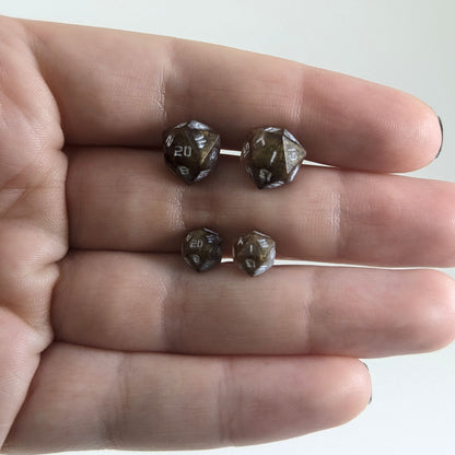 Two pairs of handmade d20 earrings with titanium hardware that are brown and yellow tiger's eye swirls with white numbers held between the fingers of a flat hand. The larger pair is between the pointer and middle finger, the smaller between the middle and ring finger. The earrings on the left of each pair centers the 20 face and the earrings on the right of each pair centers the 1 face.