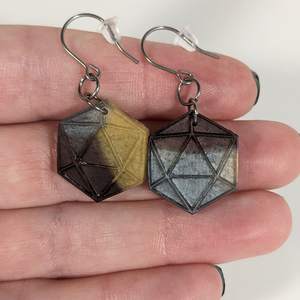 A pair of gold, maroon, and dark silver dangly, flat d20 earrings with silver color titanium and niobium hardware with bell shaped rubber backs held on on a hand on a white background.
