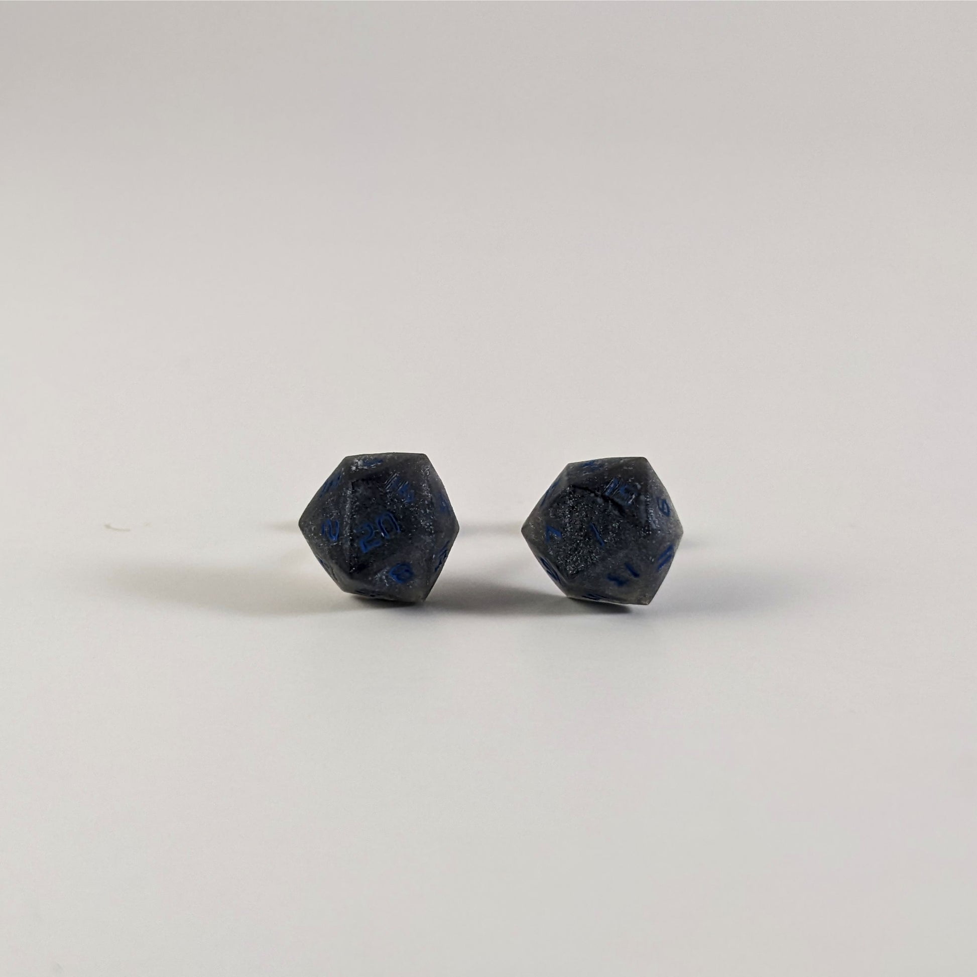 A single pair of the larger size handmade d20 earrings that are black and dark silver with blue numbers on a white background. The earring on the left centers the 20 face and the earring on the right centers the 1 face.