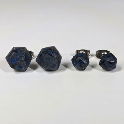 Two pairs of handmade d20 earrings that are black and dark silver with blue numbers on a white background sit in a line with the larger pair on the left and the smaller pair on the right. The earrings on the left of each pair centers the 20 face and the earrings on the right of each pair centers the 1 face.