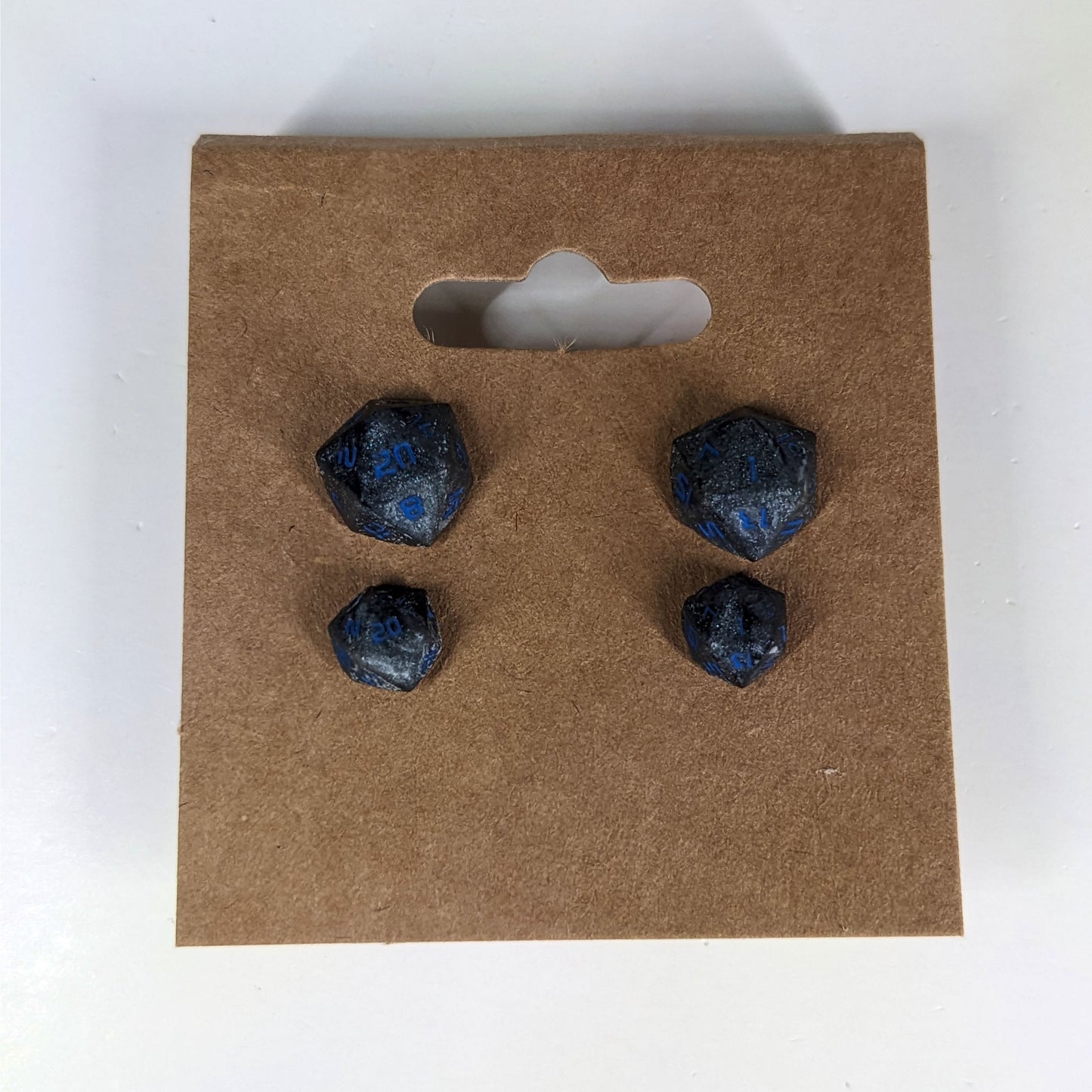 Two pairs of handmade d20 earrings that are black and dark silver with blue numbers on a kraft brown earring card. The earrings on the left of each pair centers the 20 face and the earrings on the right of each pair centers the 1 face.