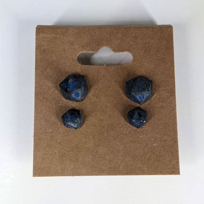Two pairs of handmade d20 earrings that are black and dark silver with blue numbers on a kraft brown earring card. The earrings on the left of each pair centers the 20 face and the earrings on the right of each pair centers the 1 face.