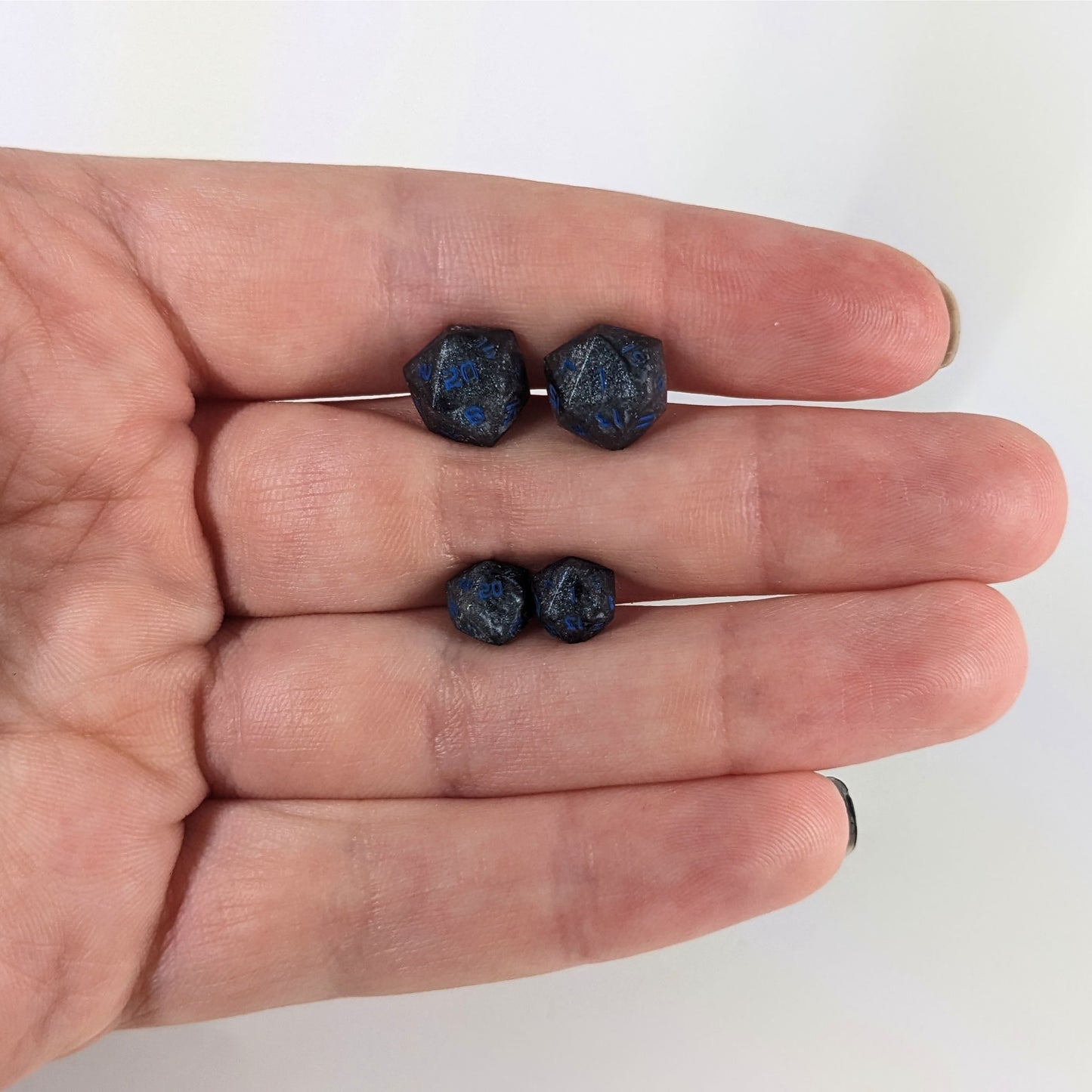 Two pairs of handmade d20 earrings that are black and dark silver with blue numbers held between the fingers of a flat hand. The larger pair is between the pointer and middle finger, the smaller between the middle and ring finger. The earrings on the left of each pair centers the 20 face and the earrings on the right of each pair centers the 1 face.