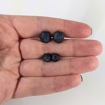Two pairs of handmade d20 earrings that are black and dark silver with blue numbers held between the fingers of a flat hand. The larger pair is between the pointer and middle finger, the smaller between the middle and ring finger. The earrings on the left of each pair centers the 20 face and the earrings on the right of each pair centers the 1 face.