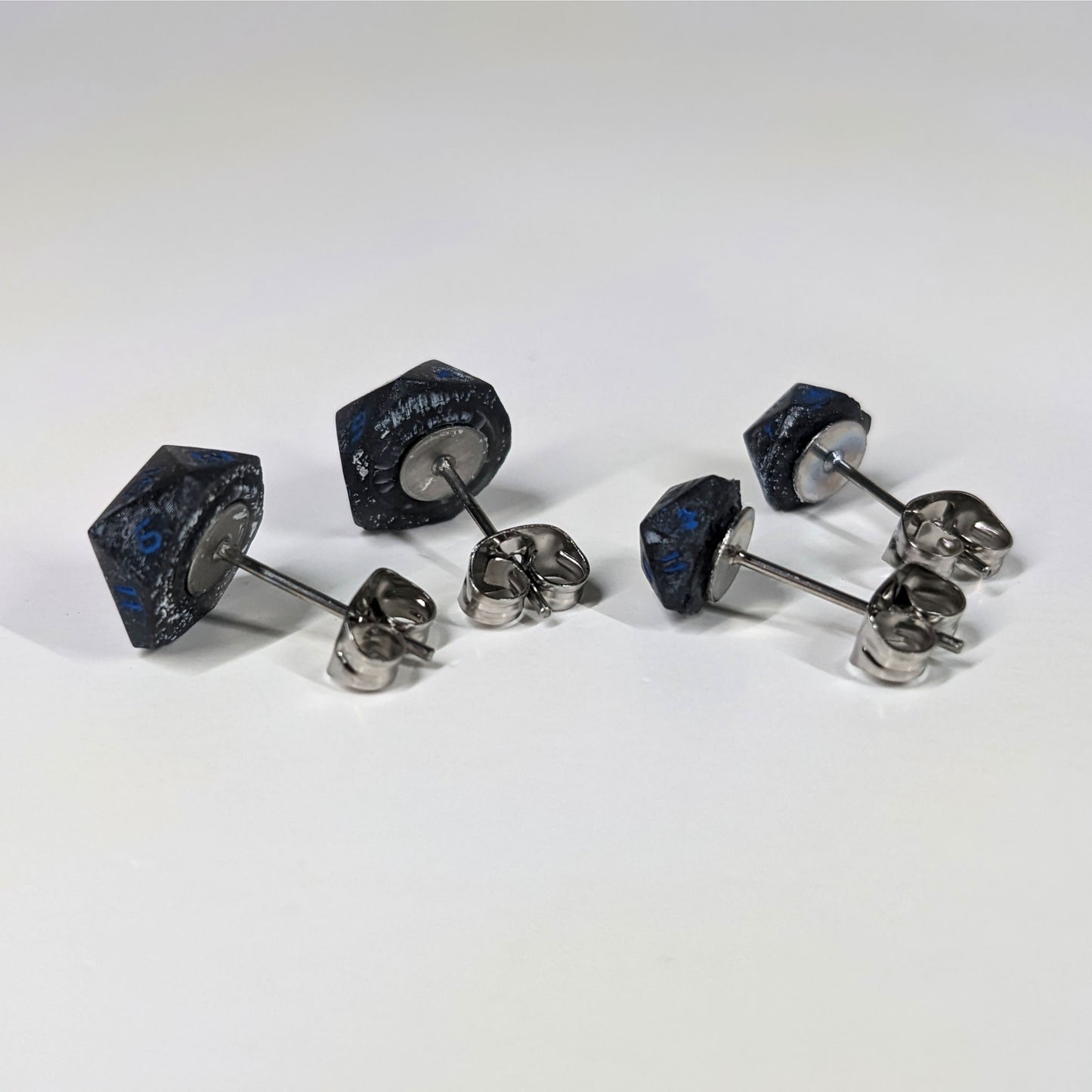 Two pairs of handmade d20 earrings that are black and dark silver with blue numbers turned to show the metal alloy hardware and butterfly backs.