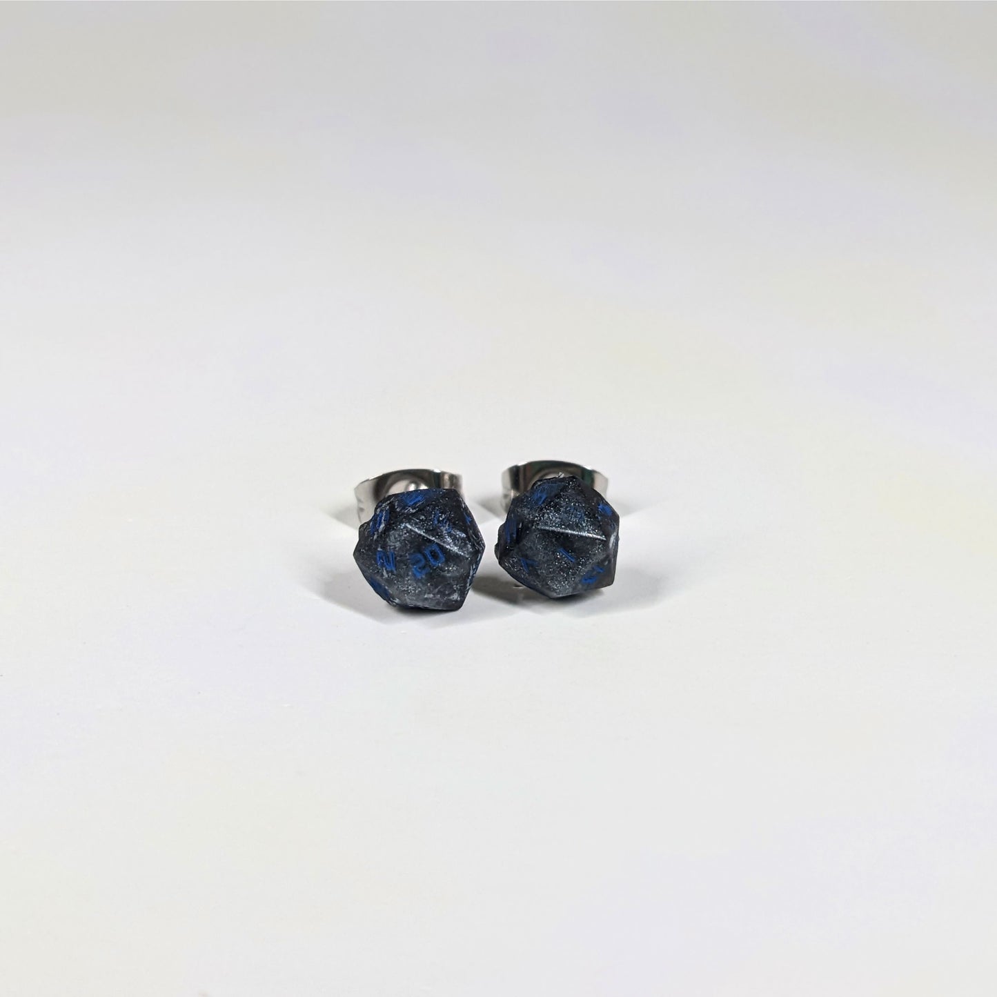 A single pair of the smaller size handmade d20 earrings that are black and dark silver with blue numbers on a white background. The earring on the left centers the 20 face and the earring on the right centers the 1 face.