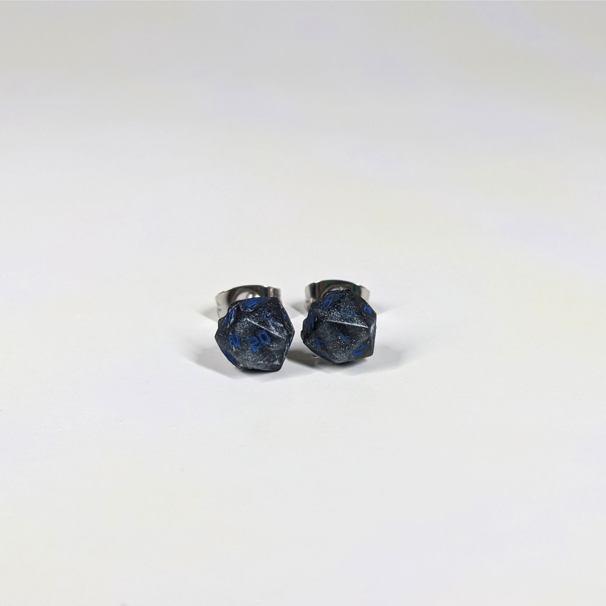 A single pair of the smaller size handmade d20 earrings that are black and dark silver with blue numbers on a white background. The earring on the left centers the 20 face and the earring on the right centers the 1 face.
