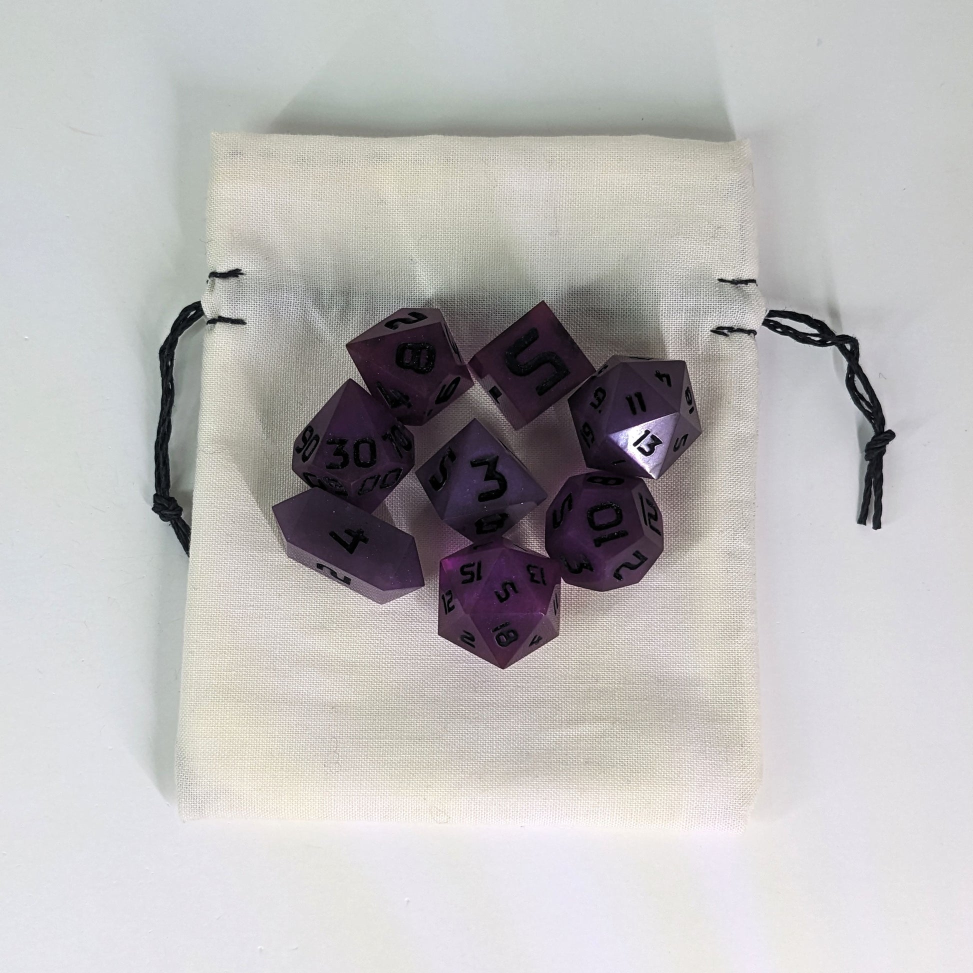 A small, handmade dice bag that is white lays flat on a white background with a set of purple dnd dice with black numbers sitting on top of it.