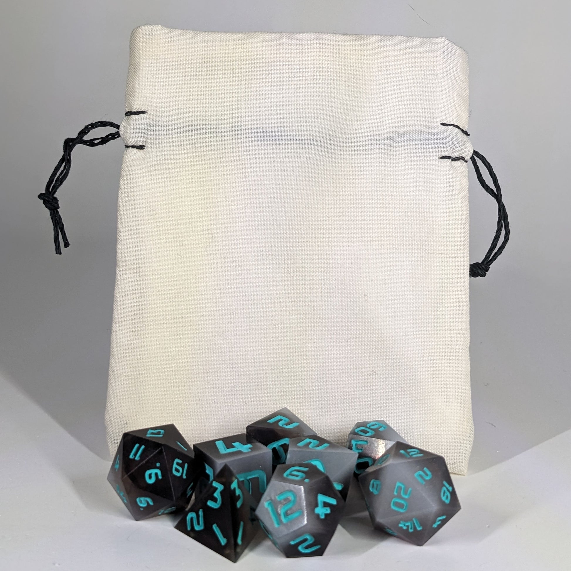A small, handmade dice bag that is white stands upright on a white background with a set of brown and white dnd dice with teal numbers sitting in front of it.