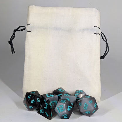 A small, handmade dice bag that is white stands upright on a white background with a set of brown and white dnd dice with teal numbers sitting in front of it.