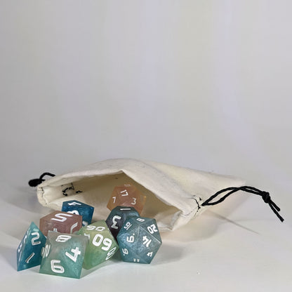 A small, handmade dice bag that is white lays open with a set of colorful, dnd dice with white numbers spilling out of it on a white background.