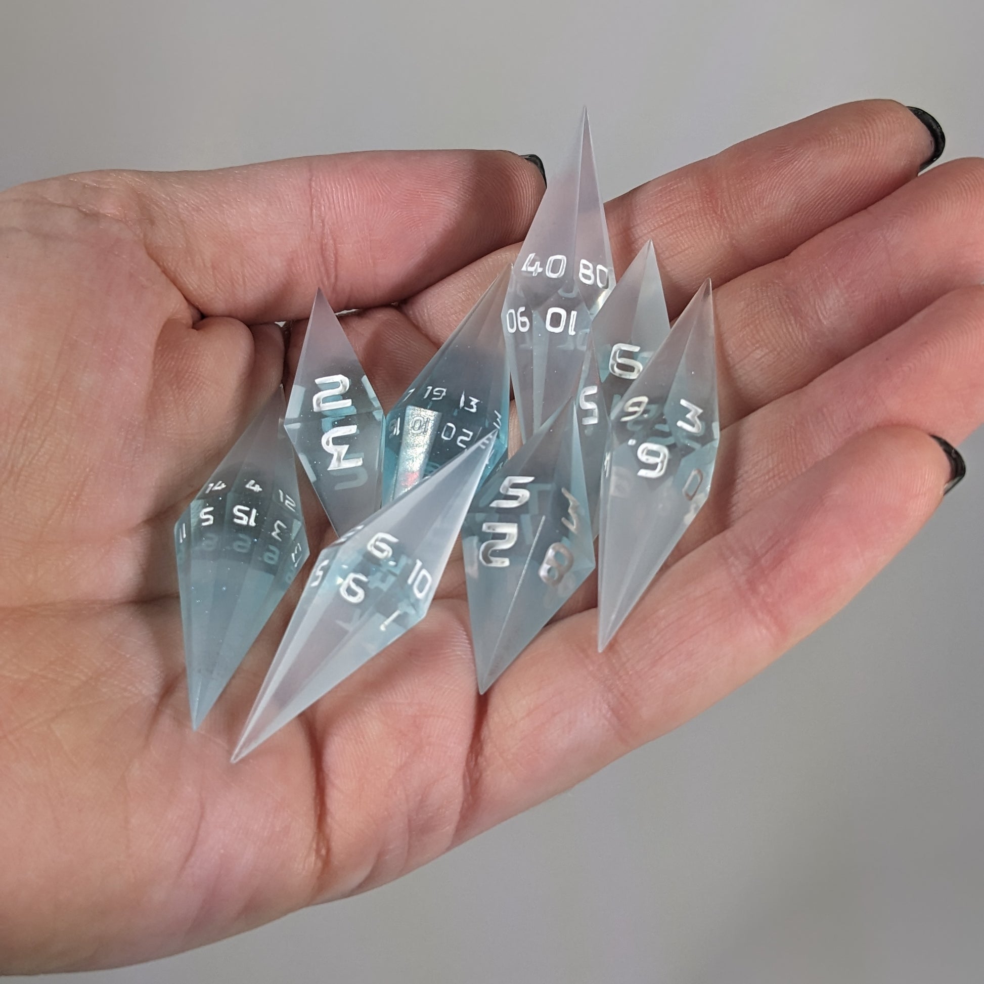 Close-up of mixed blue and white semitransparent with white numbering  artisan dice resting in a palm on a seamless white backdrop. The handmade dice feature a distinct sharp diamond shape, with 2 d20s visible among the high-quality resin dice.