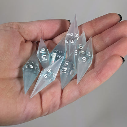 Close-up of mixed blue and white semitransparent with white numbering  artisan dice resting in a palm on a seamless white backdrop. The handmade dice feature a distinct sharp diamond shape, with 2 d20s visible among the high-quality resin dice.