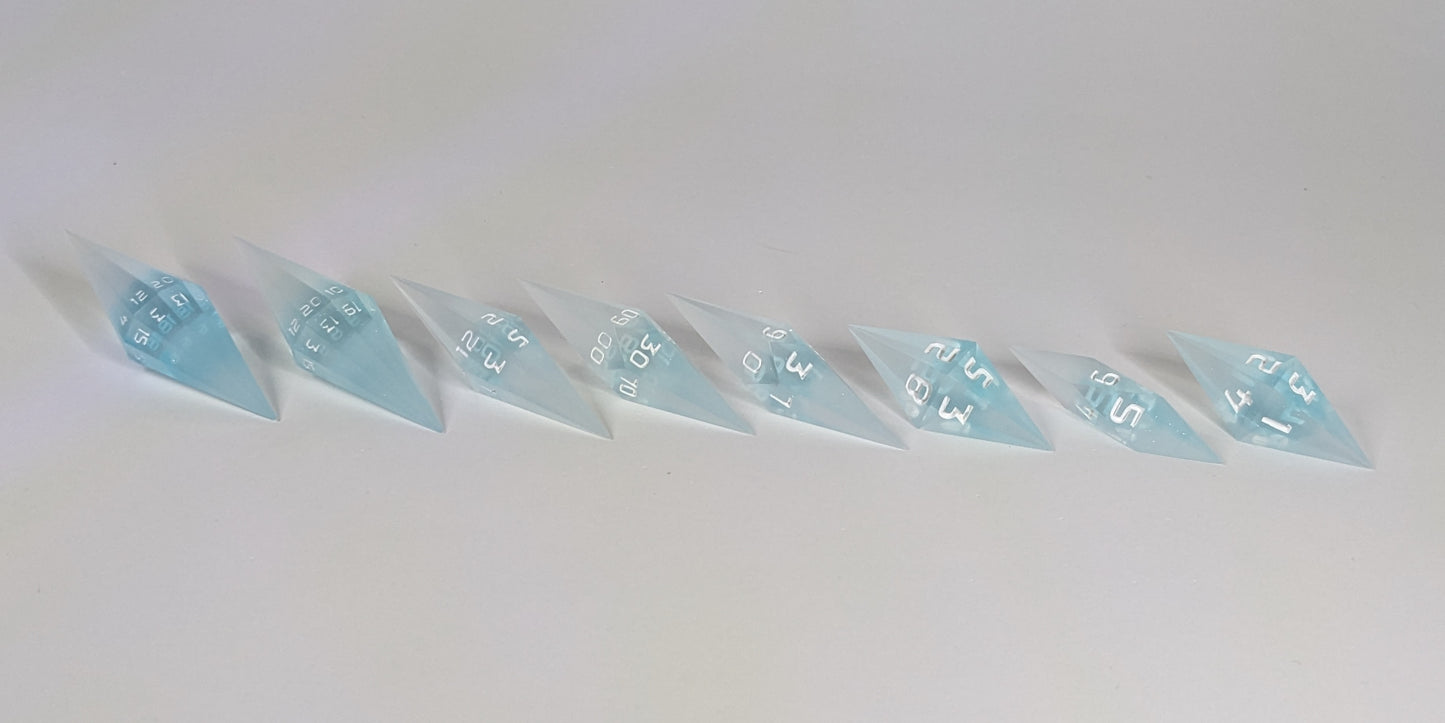 A side profile photograph of a complete set of mixed blue and white semitransparent with white numbering handmade dice arranged in a straight line. This eye-level view emphasizes the unique elongated and pointed diamond silhouette of the resin dice, showing the sharp geometry of the 2 d20s and matching artisan dice.
