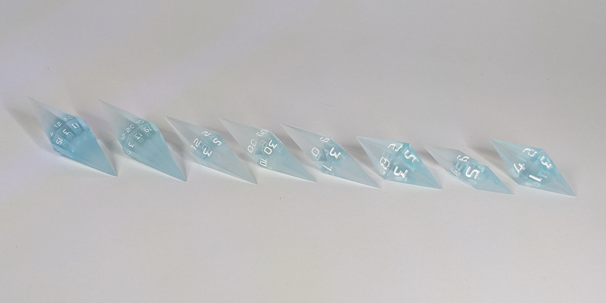 A side profile photograph of a complete set of mixed blue and white semitransparent with white numbering handmade dice arranged in a straight line. This eye-level view emphasizes the unique elongated and pointed diamond silhouette of the resin dice, showing the sharp geometry of the 2 d20s and matching artisan dice.
