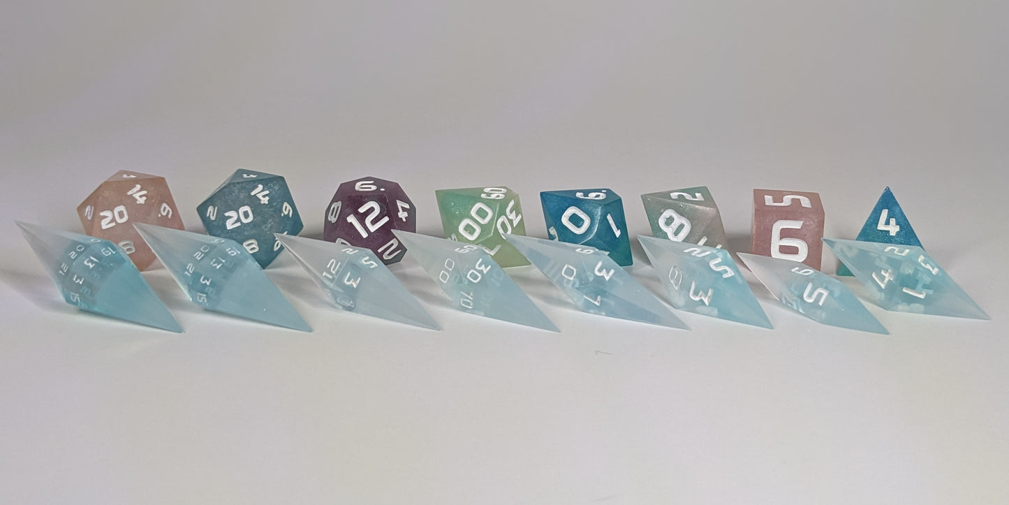 A size comparison of mixed blue and white semitransparent with white numbering long pointed diamond handmade dice with standard shapes placed behind for scale. The photo emphasizes the length of the diamond dice being much greater than a standard set as well as the diamond set being much more slender. This artisan dice set features 2 d20s and rests on a seamless white background to highlight the elongated profile of the resin dice.