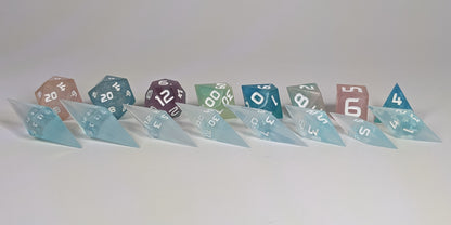 A size comparison of mixed blue and white semitransparent with white numbering long pointed diamond handmade dice with standard shapes placed behind for scale. The photo emphasizes the length of the diamond dice being much greater than a standard set as well as the diamond set being much more slender. This artisan dice set features 2 d20s and rests on a seamless white background to highlight the elongated profile of the resin dice.