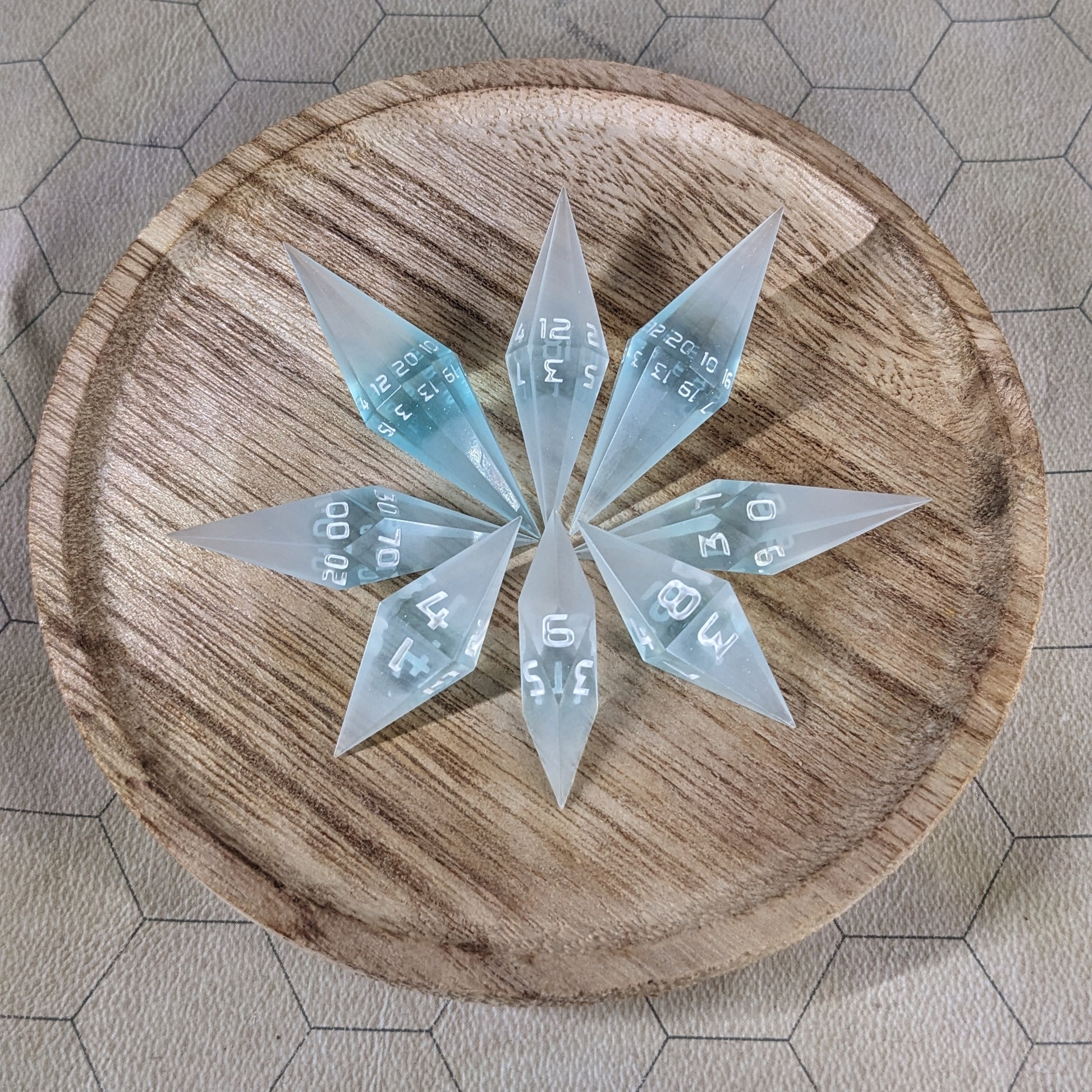 A set of mixed blue and white semitransparent with white numbering handmade dice arranged in a wooden circle organizer. The resin dice feature a unique pointed diamond shape with the tips facing outward, highlighting the sharp edges of the two d20 and other artisan dice in the collection. 