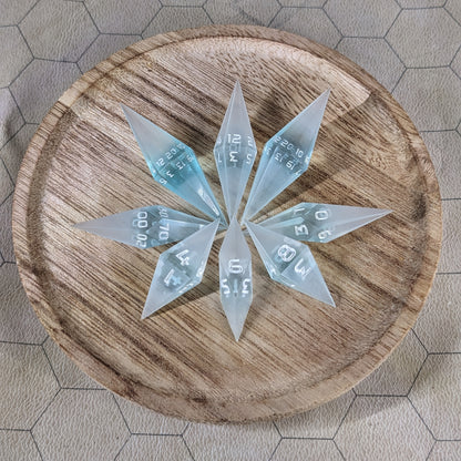 A set of mixed blue and white semitransparent with white numbering handmade dice arranged in a wooden circle organizer. The resin dice feature a unique pointed diamond shape with the tips facing outward, highlighting the sharp edges of the two d20 and other artisan dice in the collection. 