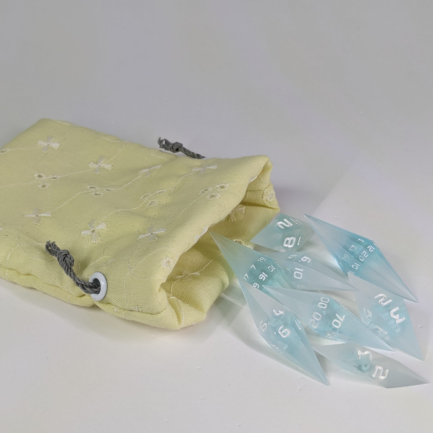Mixed blue and white semitransparent with white numbering diamond handmade dice spilling out of a yellow patterned drawstring bag on a white background. The set features 2 d20s and matching resin dice, showcasing the unique shape of these artisan dice.