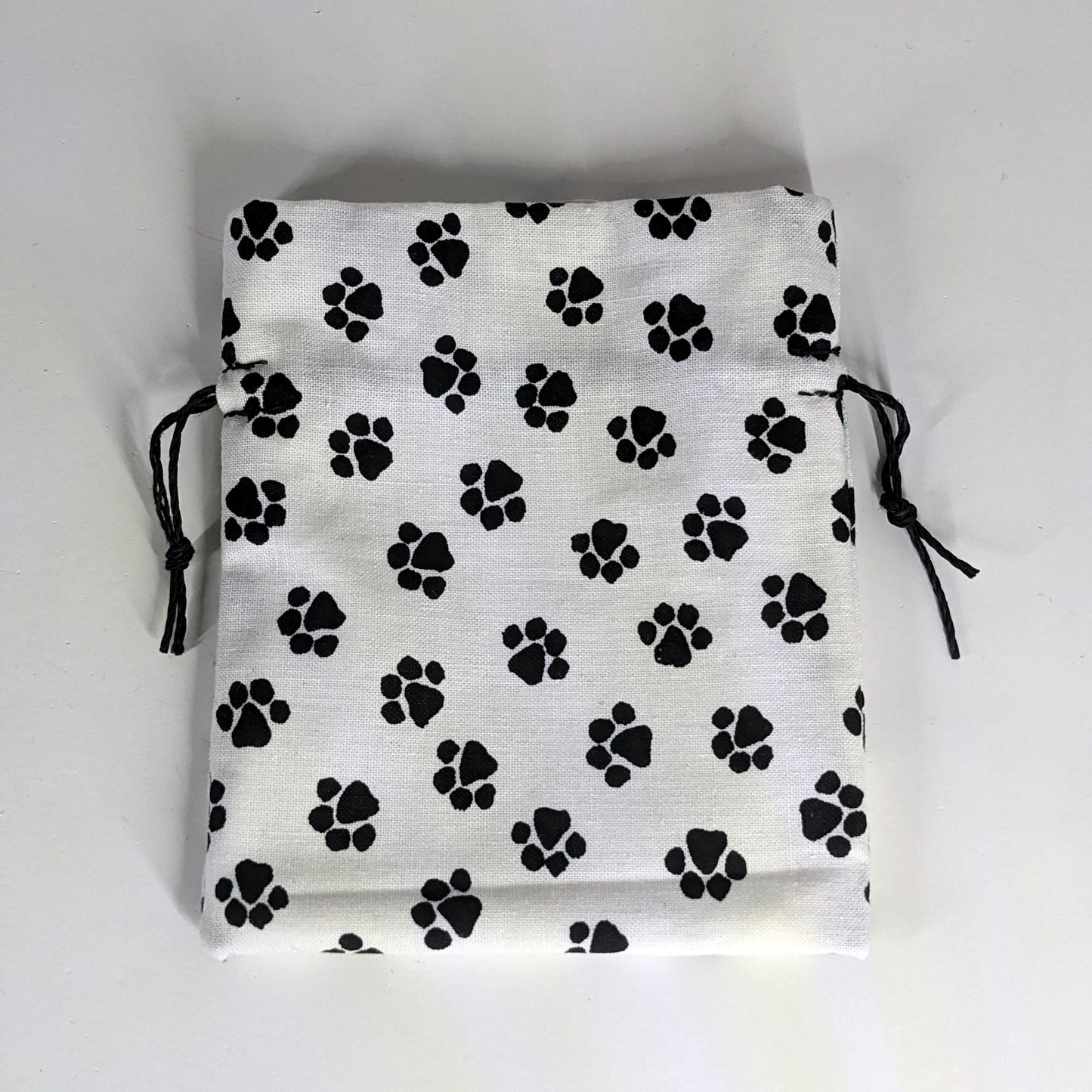 A small, handmade dice bag that is white with a black paw print pattern lays flat on a white background.