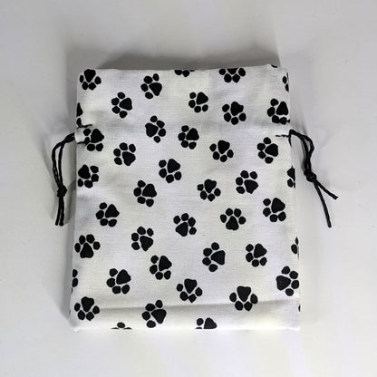 A small, handmade dice bag that is white with a black paw print pattern lays flat on a white background.