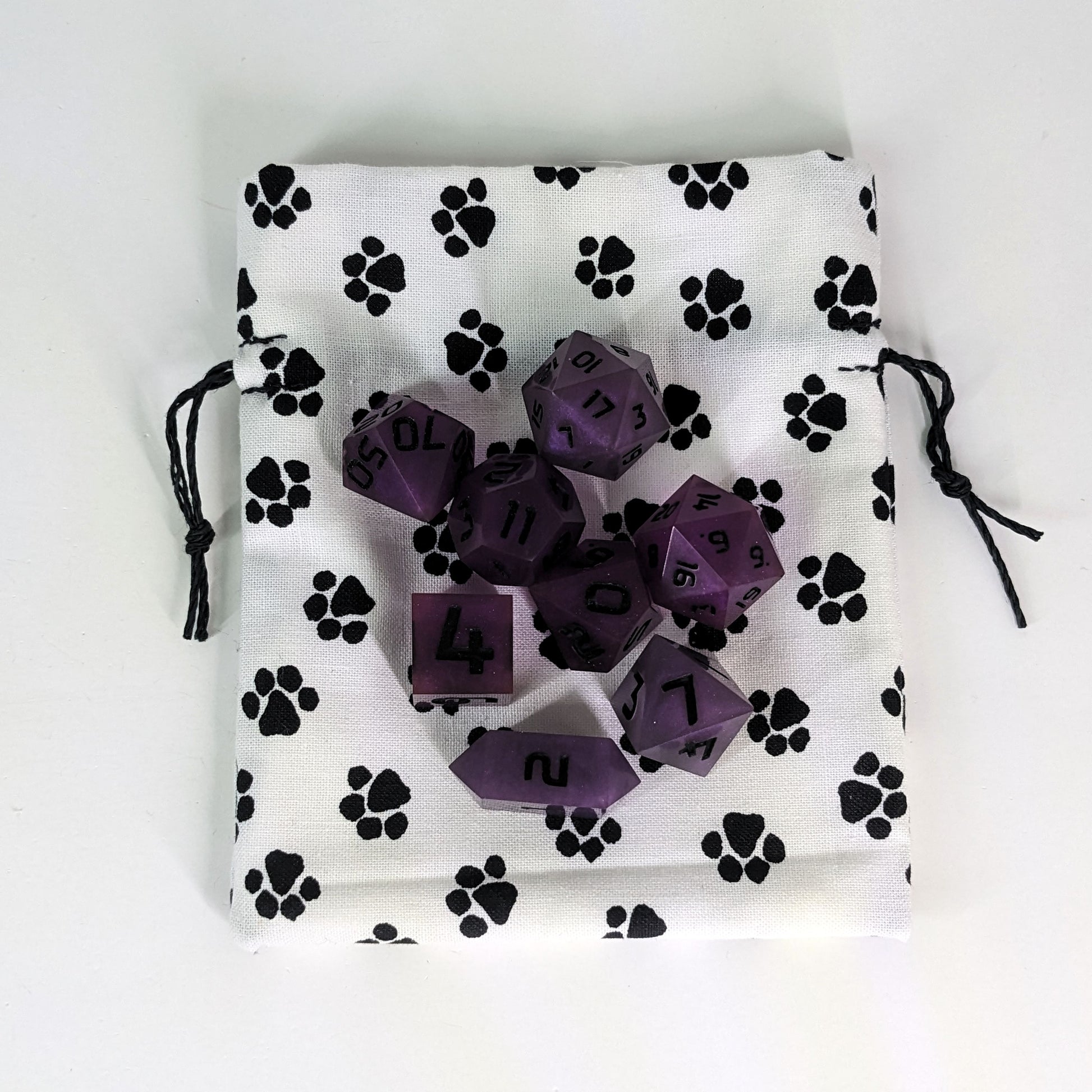 A small, handmade dice bag that is white with a black paw print pattern lays flat on a white background with a set of purple dnd dice sitting on top of the bag.