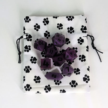 A small, handmade dice bag that is white with a black paw print pattern lays flat on a white background with a set of purple dnd dice sitting on top of the bag.