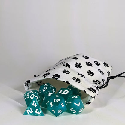 A small, handmade dice bag that is white with a black paw print pattern lays open with a set of teal, polyhedral dice spilling out of it on a white background.