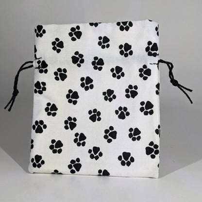 A small, handmade dice bag that is white with a black paw print pattern stands upright on a white background.