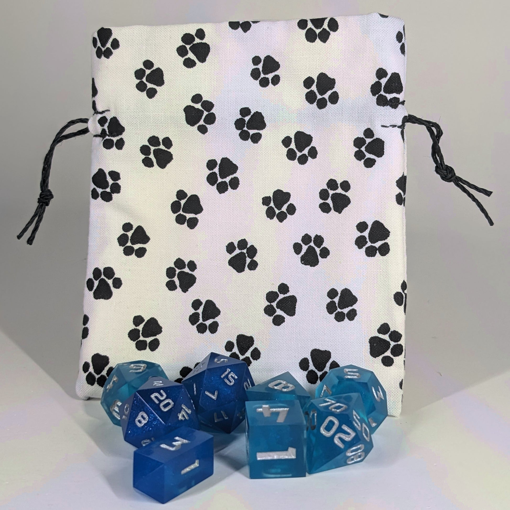 A small, handmade dice bag that is white with a black paw print pattern stands upright on a white background with a set of blue dnd dice sitting in front of the bag.