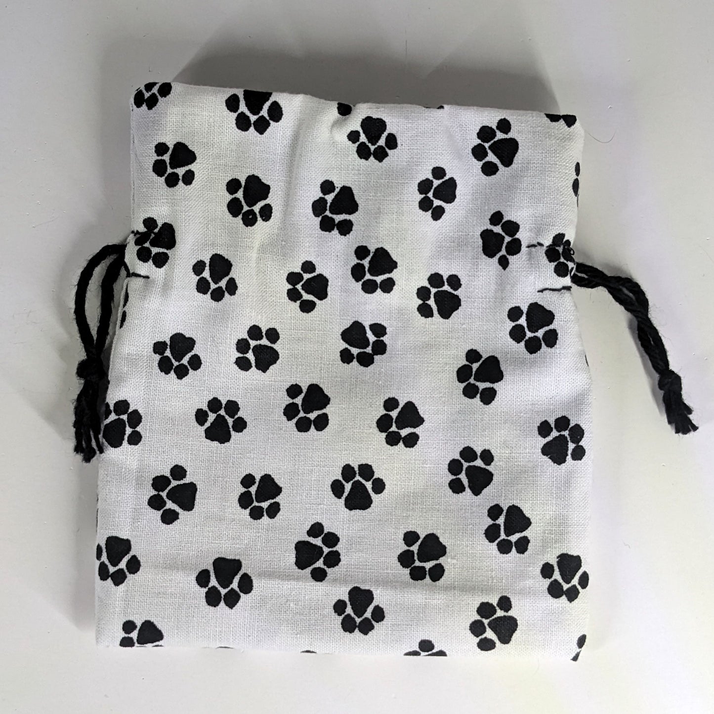 A small, handmade dice bag that is white with a black paw print lays flat on a white background.